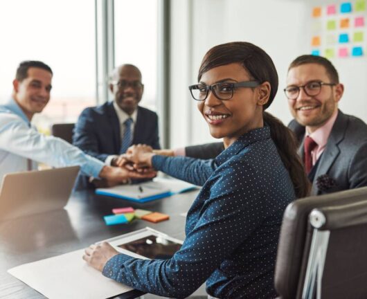 Successful African American team leader turning to smile at the camera as her multiracial team of executives links hands across the table